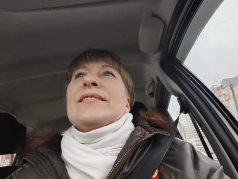 Portrait of adult cute funny blonde woman in warm clothes taking a selfie inside the car and driving it. The concept of pleasant trip in spring, autumn, winter