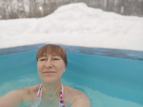 Adult mature woman takes selfie in a pool with warm hot termal mineral water in winter and white snow around. Wellness center and the concept of health care. Travel, recreation, medicine and rest