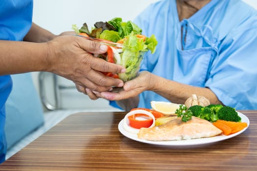 Asian elderly woman patient eating Salmon steak breakfast with vegetable healthy food while sitting and hungry on bed in hospital.