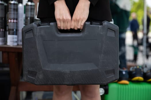 A woman's hands hold a black plastic suitcase with power tools by the handle. Scene taken on a sunny day outside.