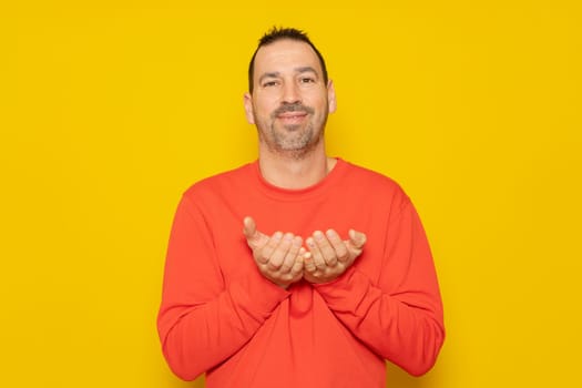 Bearded Hispanic man dressed in a red jumper offering something in his hands, has a friendly and receptive demeanor. Isolated on yellow background.