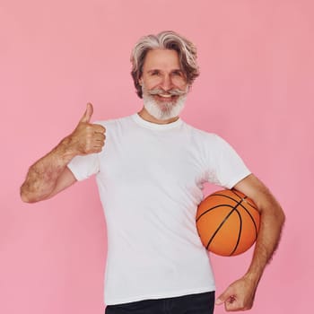 With sport ball in hands. Stylish modern senior man with gray hair and beard is indoors.