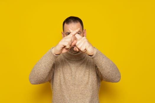 Bearded hispanic man in beige turtleneck shows hands to stop timeout, X fingers. Isolated over yellow background