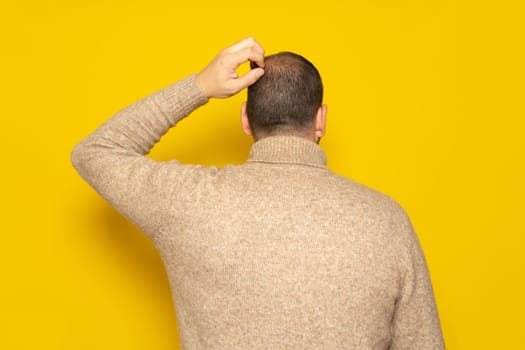 Hispanic man wearing a beige turtleneck scratching his head with his back to the camera in concept of doubt or worry, isolated over yellow background