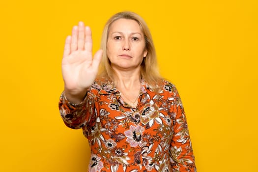 Caucasian blonde woman in a patterned dress raising her palm in a stop sign, isolated over yellow background