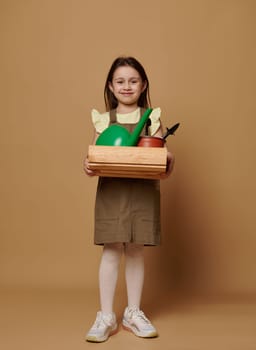 Full-length portrait of a conscious Caucasian child girl holding a wooden box with garden tools, isolated on beige background. Kids. Agricultural hobby. Earth Day. Environmental conservation concept