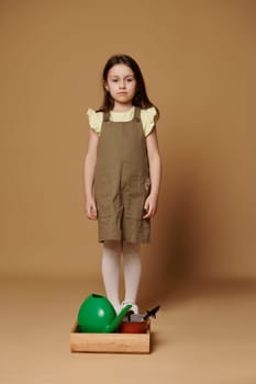 Serious conscious Caucasian 5 y.o child girl standing over beige background, near a wooden crate with garden tools and watering can, looking confidently at camera. People and nature concept. Earth Day