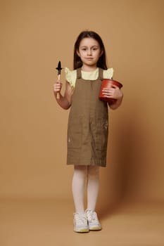 Caucasian happy little child girl smiling broadly, looking at camera, holding a flower pot, garden shovel and rake, isolated over beige background. Full-length of a cute kid dreaming becoming gardener