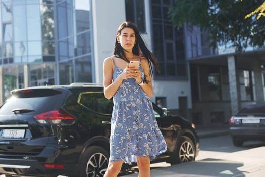 Young woman in casual blue dress and with smartphone in hands standing near black car in the city.