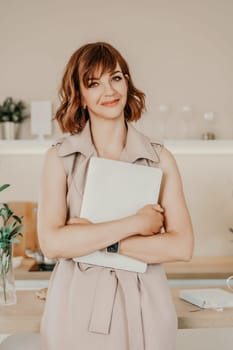 Brunette macbook. Portrait of a woman, she holds a mabuk in her hands and looks at the camera, wearing a beige dress on a beige background