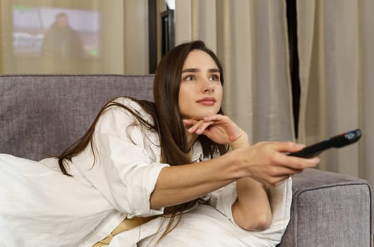 Positive young woman sitting on the sofa indoors at home and watching TV with remote control on the sofa.