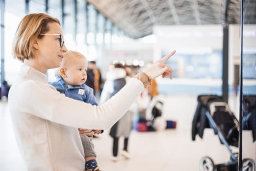 Mother traveling with child, holding his infant baby boy at airport terminal, checking flight schedule, waiting to board a plane. Travel with kids concept