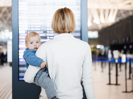 Mother traveling with child, holding his infant baby boy at airport terminal, checking flight schedule, waiting to board a plane. Travel with kids concept