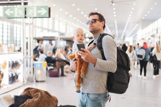 Father traveling with child, holding his infant baby boy at airport terminal, checking flight schedule, waiting to board a plane. Travel with kids concept