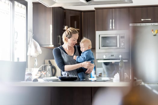 Happy mother and little infant baby boy making pancakes for breakfast together in domestic kitchen. Family, lifestyle, domestic life, food, healthy eating and people concept