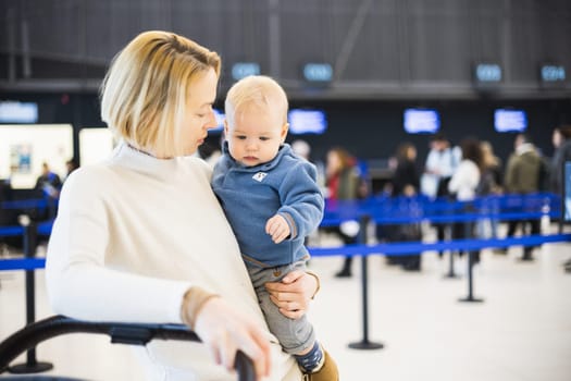 Motherat travelling with his infant baby boy child, walking, pushing baby stroller and luggage cart at airport terminal station. Travel with child concept