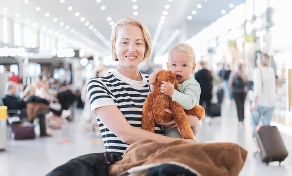 Mother traveling with child, holding his infant baby boy at airport terminal waiting to board a plane. Travel with kids concept