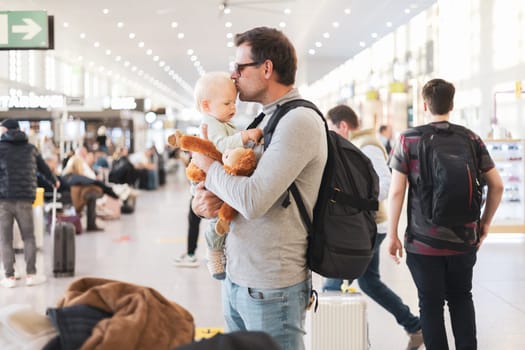 Father traveling with child, holding and kissing his infant baby boy at airport terminal waiting to board a plane. Travel with kids concept