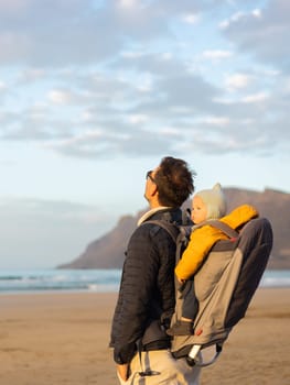 Father rising hands to the sky while enjoying pure nature carrying his infant baby boy son in backpack on windy sandy beach of Famara, Lanzarote island, Spain at sunset. Family travel concept