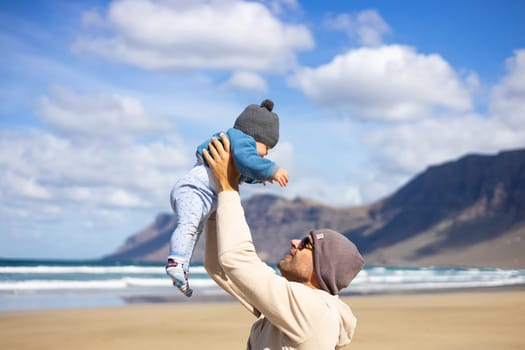 Father enjoying pure nature holding and playing with his infant baby boy son in on windy sandy beach of Famara, Lanzarote island, Spain. Family travel and parenting concept