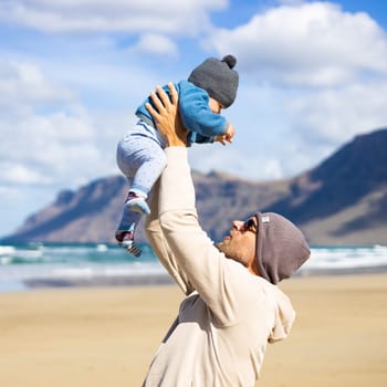 Father enjoying pure nature holding and playing with his infant baby boy son in on windy sandy beach of Famara, Lanzarote island, Spain. Family travel and parenting concept