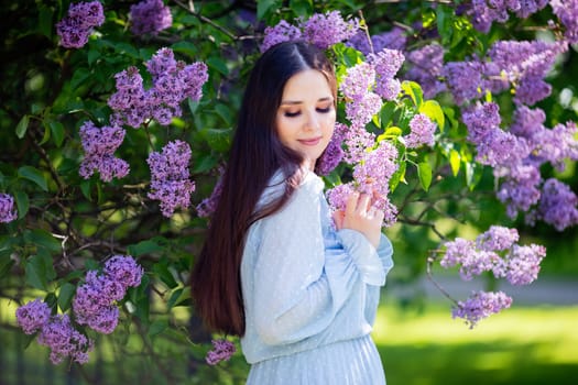 A beautiful girl in light blue dress, stands next to a flowering lilac bush in the park. Looking down. Close up. Copy space