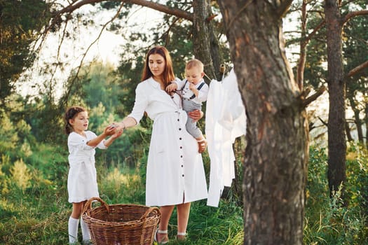Housewife drying clothes. Young mother with her little daughter and son is outdoors in the forest. Beautiful sunshine.