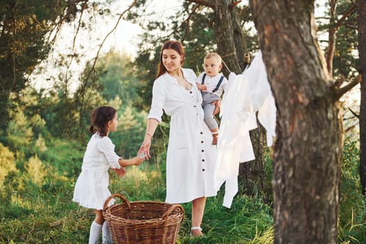 Housewife drying clothes. Young mother with her little daughter and son is outdoors in the forest. Beautiful sunshine.