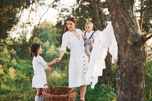 Housewife drying clothes. Young mother with her little daughter and son is outdoors in the forest. Beautiful sunshine.