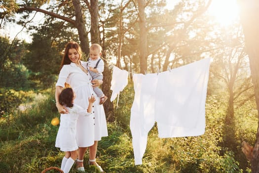 White clothes hanging on the rope to dry. Young mother with her little daughter and son is outdoors in the forest. Beautiful sunshine.