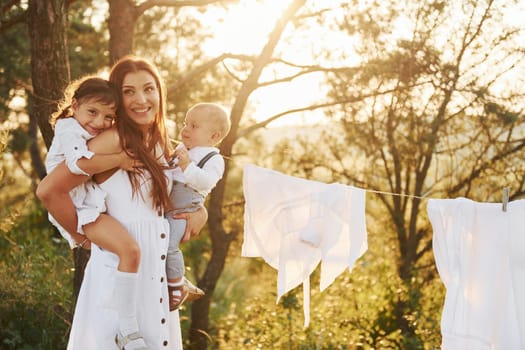 White clothes hanging on the rope to dry. Young mother with her little daughter and son is outdoors in the forest. Beautiful sunshine.