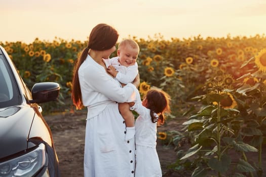 Near modern black car. Young mother with her little son and daughter is outdoors in the agricultural field. Beautiful sunshine.