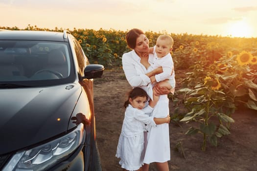 Near modern black car. Young mother with her little son and daughter is outdoors in the agricultural field. Beautiful sunshine.
