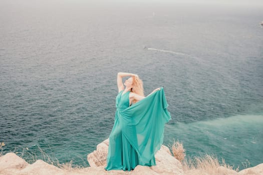 Side view a Young beautiful sensual woman in a mint long dress posing on a volcanic rock high above the sea during sunset. Girl on the nature on overcast sky background. Fashion photo