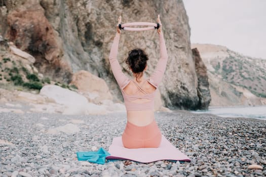 Middle aged well looking woman with black hair doing Pilates with the ring on the yoga mat near the sea on the pebble beach. Female fitness yoga concept. Healthy lifestyle, harmony and meditation.