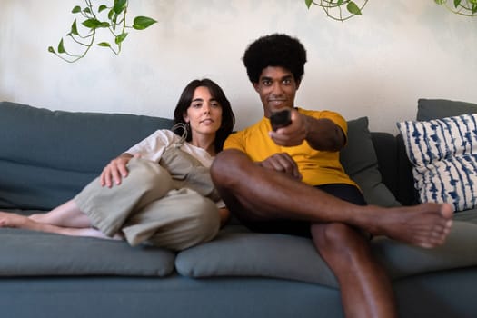 Young multiracial couple watch TV at home. African American man changes channel with remote control. Lifestyle concept.