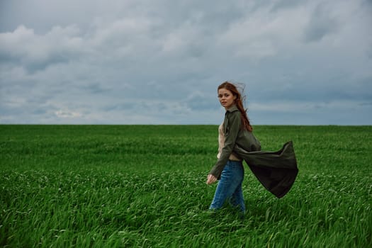 A woman stands in a green field in a raincoat and looks at the camera. Strong wind, flying hair, freedom and harmony with nature. High quality photo