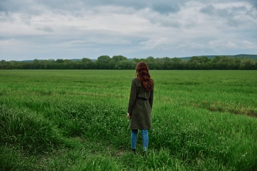 a woman with beautiful, long, red hair stands with her back to the camera in a green field in rainy, spring weather in a long raincoat. High quality photo