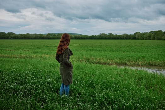 a woman with beautiful, long, red hair stands with her back to the camera in a green field in rainy, spring weather in a long raincoat. High quality photo