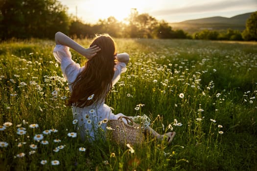 a red-haired woman in a light dress sits with her back to the camera in a field of daisies straightening her hair with her hands against the backdrop of the setting sun. High quality photo