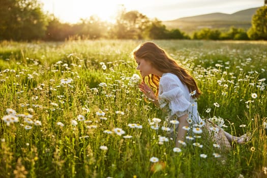 a beautiful woman in a light dress sits in a field of daisies against the backdrop of the setting sun and inhales their fragrance. High quality photo