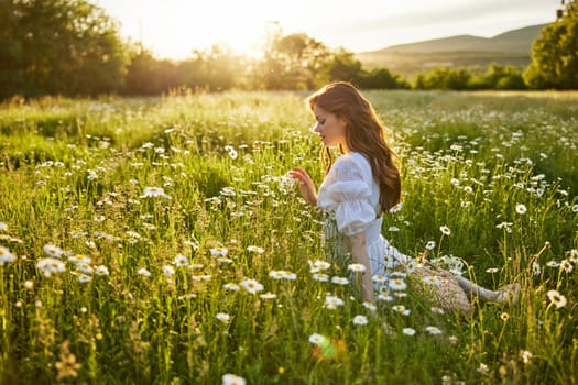 a beautiful woman in a light dress sits in a field of daisies against the backdrop of the setting sun and inhales their fragrance. High quality photo