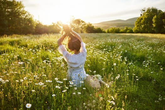 a red-haired woman in a light dress sits with her back to the camera in a field of daisies straightening her hair with her hands against the backdrop of the setting sun. High quality photo