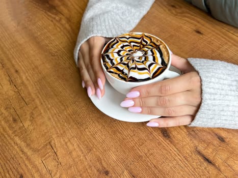 Girl's hands hold white cup of coffee with spider web pattern on top of foam, against background of wooden table, close-up, top view