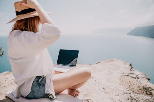 Successful business woman in yellow hat working on laptop by the sea. Pretty lady typing on computer at summer day outdoors. Freelance, travel and holidays concept.