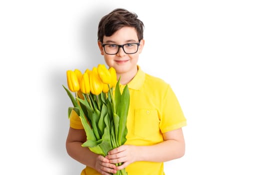 A happy cute boy in glasses and a yellow T-shirt holds a bouquet of yellow tulips, stands on a white background, isolated. Close up. Copy space.