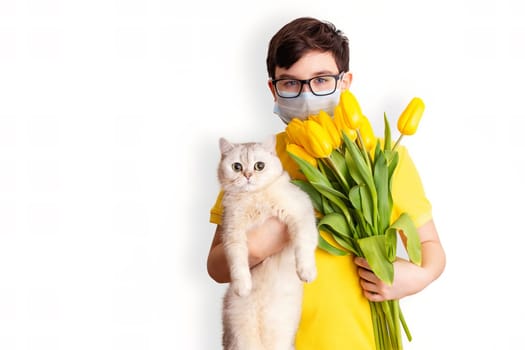 Portrait of a dark-haired boy with glasses and a medical mask, wearing a yellow T-shirt, holding a white cat and a bouquet of yellow tulips , on a white background. Close up. Copy space