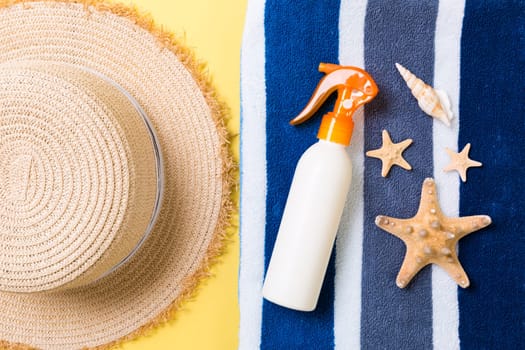 Beach flat lay accessories with copy space. Striped blue and white towel, seashells, staw sunhat and a bottle of sunblock on yellow background. Summer holiday concept.