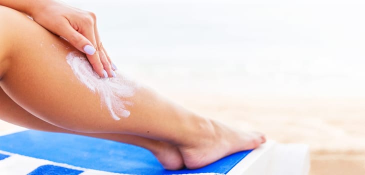 Young woman is having rest at the beach and protects her skin applying sunblock on her leg.