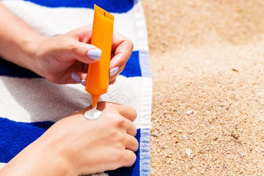 Attractive woman is lying on a striped towel at the beach and squeezing sunblock from a tube on her hand.